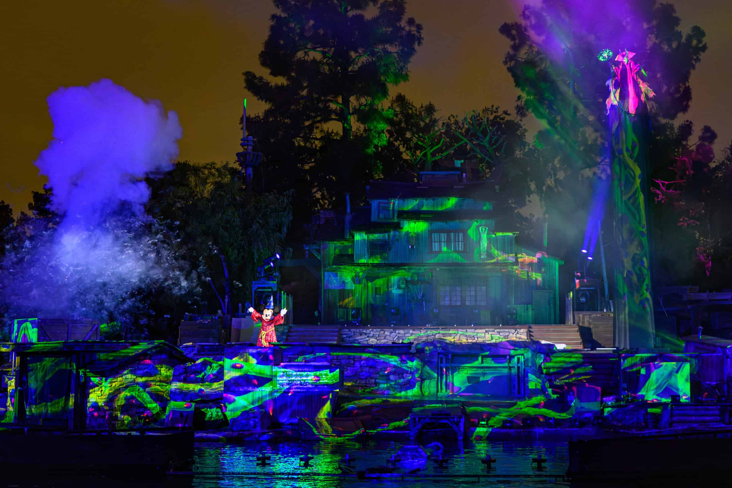 A vibrant nighttime scene from a live Fantasmic! show at Disney. A central character in red stands surrounded by colorful, glowing projections and special effects, including smoke and lights, with the silhouette of a dragon emerging against a backdrop of trees and a large building.