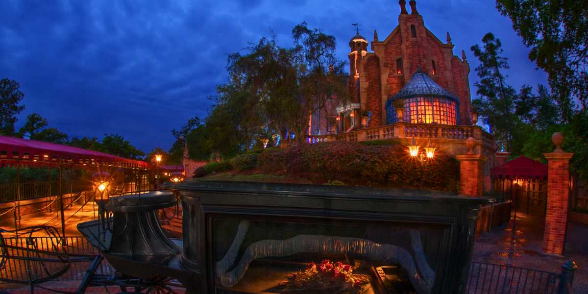 A spooky, Gothic-style haunted mansion illuminated at night with warm yellow lights. A horse-drawn hearse with no horse is in the foreground, with a dark, cloudy sky adding to the eerie atmosphere. Trees and greenery surround the mansion, enhancing the haunted mood.