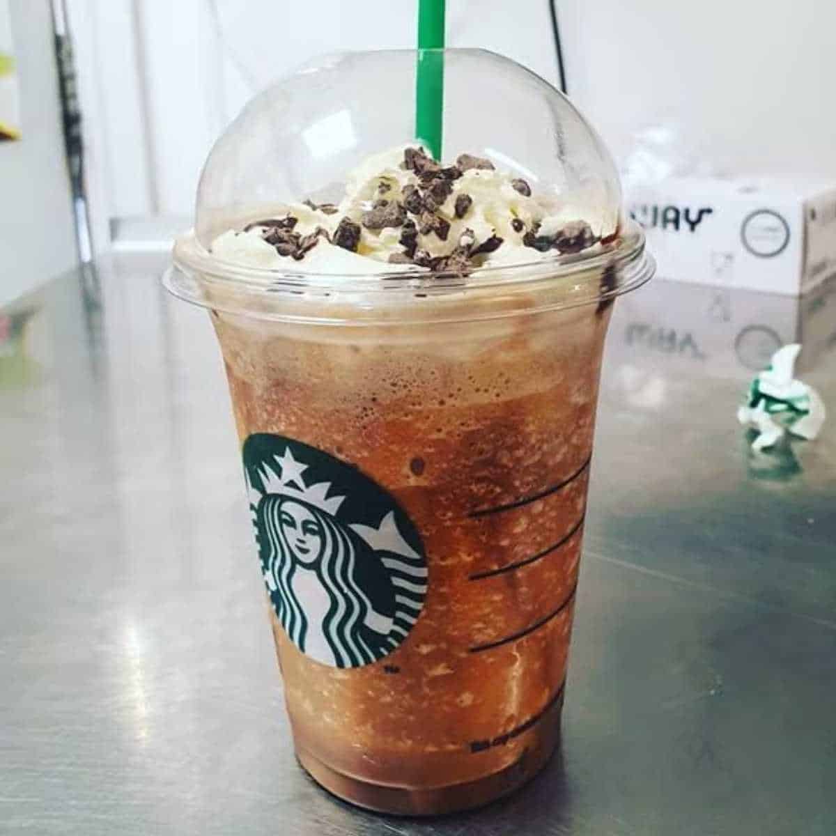 A Starbucks Jack Skellington Frappuccino topped with whipped cream and chocolate sprinkles in a clear plastic cup with a green straw on a shiny countertop. The beverage has a blended, frosty appearance. Boxes and items are blurred in the background.