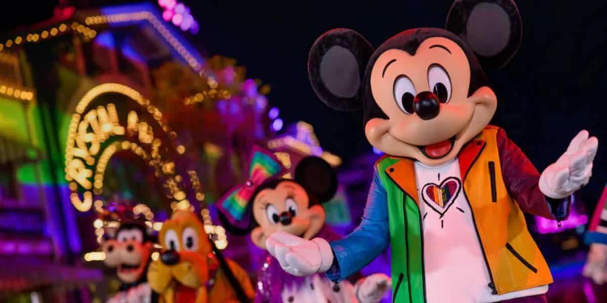 Mickey Mouse, wearing a colorful rainbow jacket from the Disney Pride merch collection, waves alongside Minnie Mouse, Goofy, and Pluto at a brightly lit, festive nighttime event.