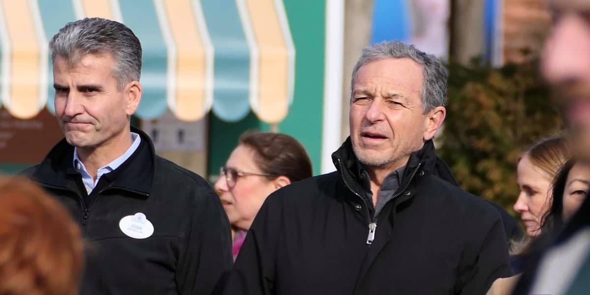 Group of people standing outdoors, including two men in the foreground wearing black jackets. The man on the left, who has a Disney Executive name tag and short grey hair, stands beside another man with short grey hair looking slightly upward. In the background is a striped awning.