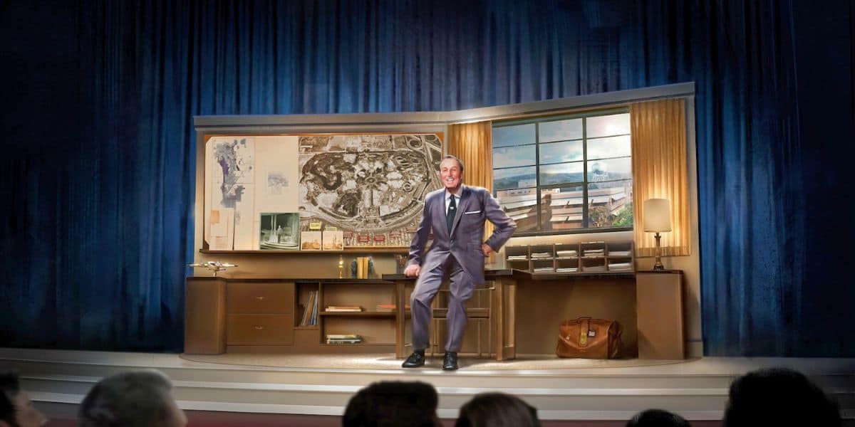 Walt Disney on stage in a suit smiling and leaning up against a desk as he presents to an audience of guests at Disneyland.