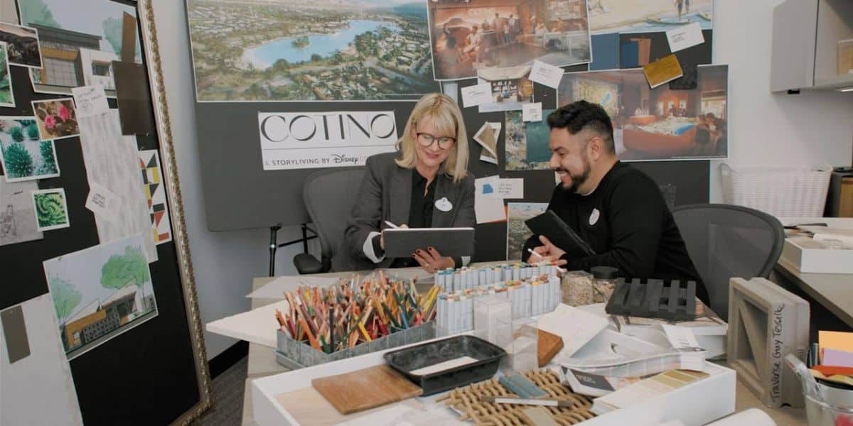 Two people sit at a desk covered with art supplies, swatches, and design materials, reviewing documents. Behind them are mood boards and images labeled "COTINO" and "A Storyliving by Disney.