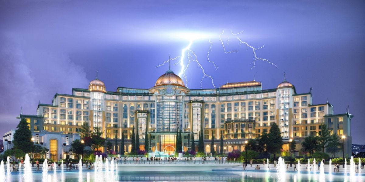 A grand, brightly lit building with domes and columns stands at night, as lightning illuminates the sky above. Water fountains in the foreground reflect the building’s lights at Epic Universe at Universal Orlando Resort.