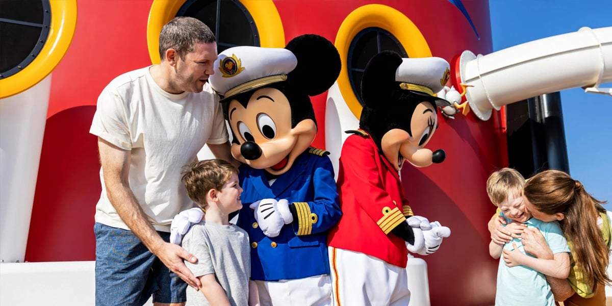 Two children and two adults interact with Mickey and Minnie Mouse in sailor outfits, standing before a colorful cruise ship backdrop under a clear blue sky, celebrating the Disney Fantasy's historic debut in Palma de Mallorca.