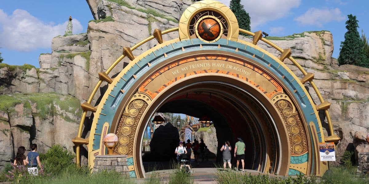 A large, ornate archway with a golden and bronze design marks the entrance to the Epic Universe theme park area. People walk through the arch, set against rocky terrain and lush landscaping under a blue sky.