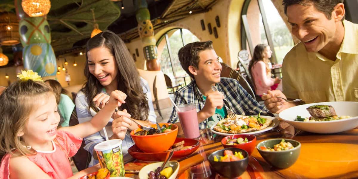 A family of four, consisting of two adults and two children, enjoys a meal together at a colorful and lively restaurant. The table is filled with various dishes, and everyone is smiling and engaged in conversation, creating a cheerful and happy atmosphere.