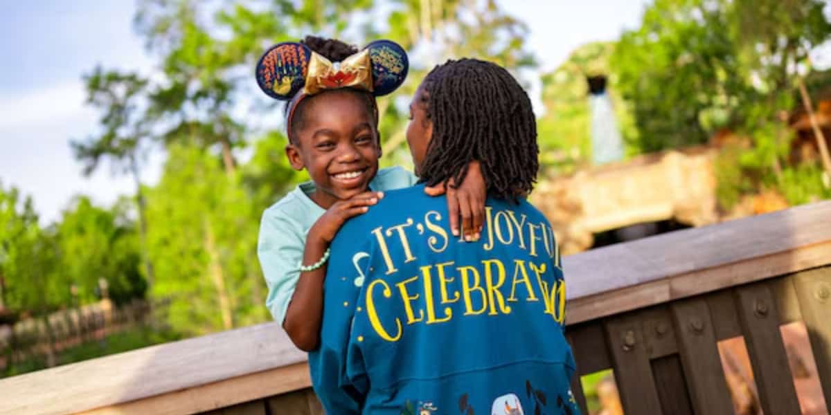 A young girl wearing Mickey Mouse ears smiles joyfully while being held up by an adult in a blue jacket with "It's Joyful Celebr..." written on the back. They're outdoors at Disney World, surrounded by trees on a sunny day.