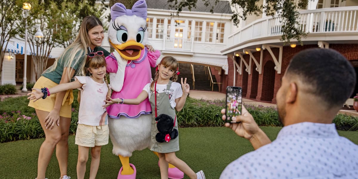 A family poses with a person dressed as Daisy Duck at an outdoor location. Two young girls stand on either side of Daisy, making peace signs, while a woman stands to the left, smiling. A man is taking a photo of the group with his smartphone as their Disney World summer commences.