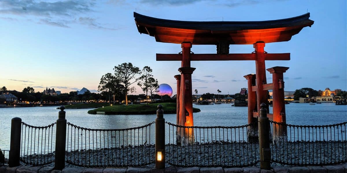 A traditional Japanese torii gate stands prominently near the edge of a lake at twilight. In the background, trees and a lit geodesic dome structure evoke scenes from Walt Disney World. The sky transitions from blue to orange as the sun sets, casting reflections on the water.