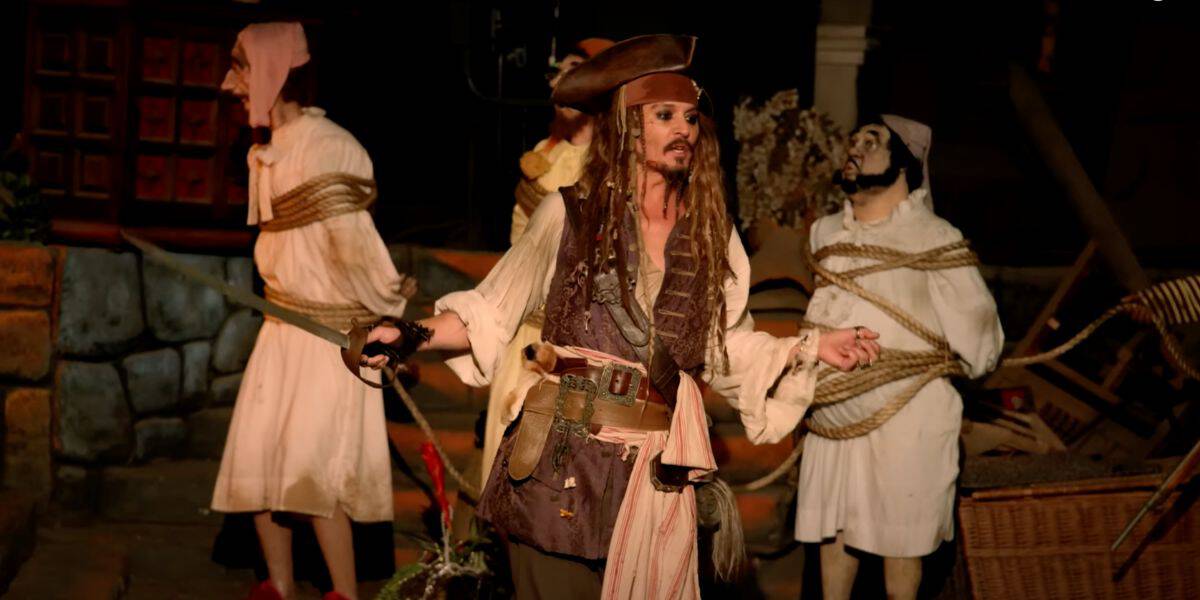 A person dressed as Jack Sparrow, with long hair and a brown hat, is standing in the foreground holding a sword. Three other people, also in pirate costumes, are tied up with ropes in the background. The scene appears to be set in a dark, dungeon-like environment.