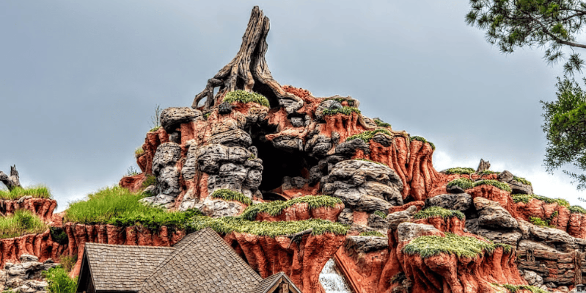 A colorful and detailed rock formation resembling a mountain, with rugged terrain, patches of greenery, and a distinct peak. The sky is overcast, adding contrast to the vibrant hues of the rocks. At its base, a wooden structure with a peaked roof hints at the Magic Kingdom's newest attraction launching next month.