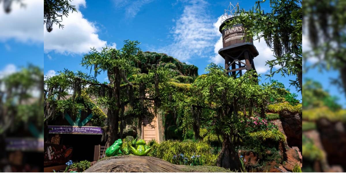 Colorful amusement park scene featuring a lush, green bayou or swamp environment with a large water tower labeled "Tiana's Foods" in the background. Floral arrangements and themed decorations are prominently displayed, with bright blue skies overhead.