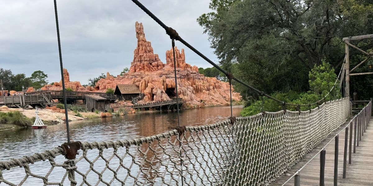 A rope suspension bridge crosses a calm river with rocky, orange mountain formations—reminiscent of Disney World changes—and wooden structures in the background, surrounded by trees and lush greenery.
