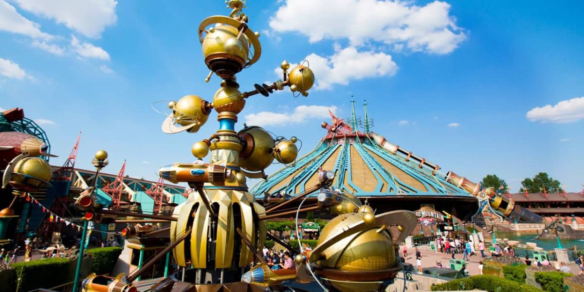 A vibrant amusement park on a sunny day featuring a futuristic, golden, planet-themed ride in the foreground. In the background, a large, conical space-themed building with colorful details stands, with several people walking around the area at this Disney Resort.