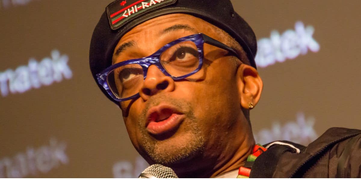 A man wearing blue glasses, a black beret with "CHI-RAQ" written on it, and a gold earring speaks into a microphone in front of a blurred background with text.