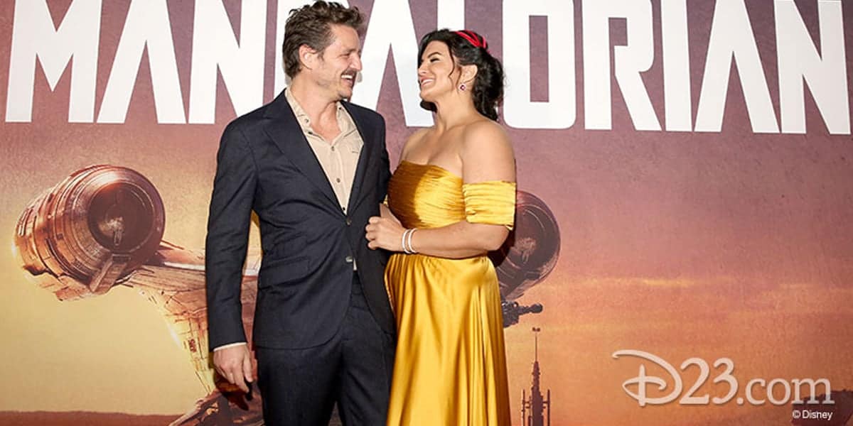 Pedro Pascal and Gina Carano at the red carpet. The man is in a dark suit, and the woman dons a yellow off-shoulder dress. Behind them, a backdrop features the word "Mandalorian" and hints of Gina Carano's recent legal battle with Disney linger in the air.