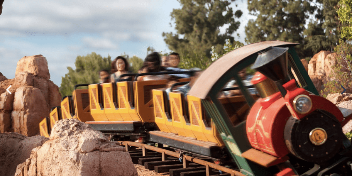 Amusement park visitors ride a colorful train-themed roller coaster. The train, with a red engine and yellow carriages, speeds along tracks amidst rocky terrain and trees. The background shows a partly cloudy sky, reminiscent of the classic Big Thunder Mountain.