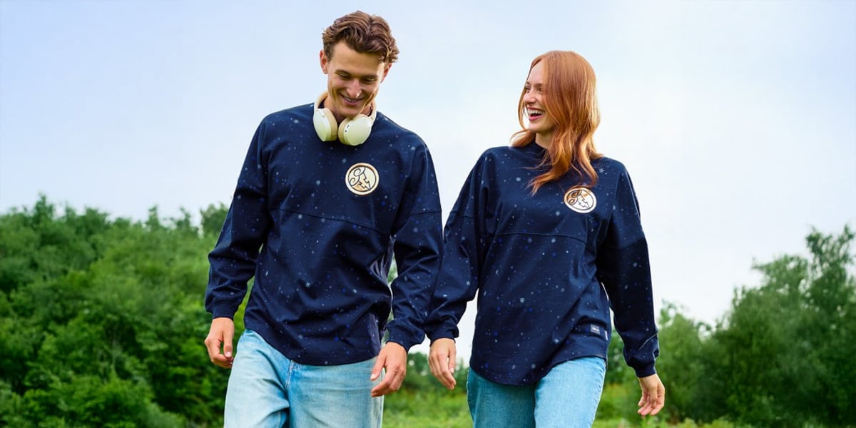 A man and woman stroll outdoors on grass, both smiling and wearing matching navy blue Spirit Jersey sweatshirts with a circular logo—channeling hero vibes. The man has headphones around his neck. Green trees and a blue sky complete the Hercules-worthy scene.