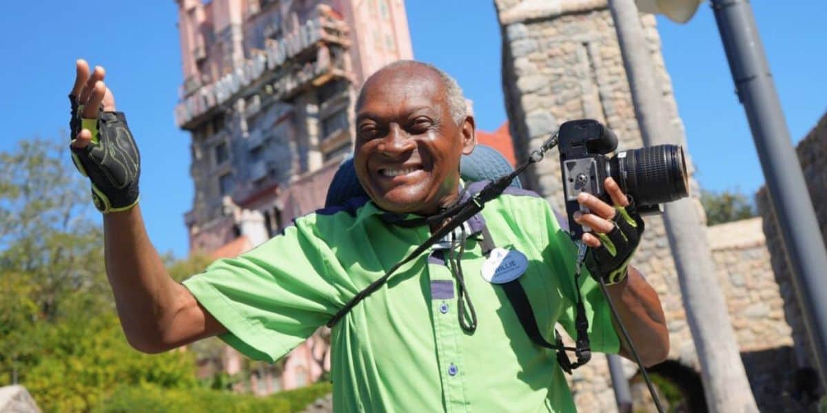A man with gray hair smiles enthusiastically while holding a camera, capturing memories at Disney. He is wearing a green shirt with a name badge and fingerless gloves, with an amusement park attraction in the background.
