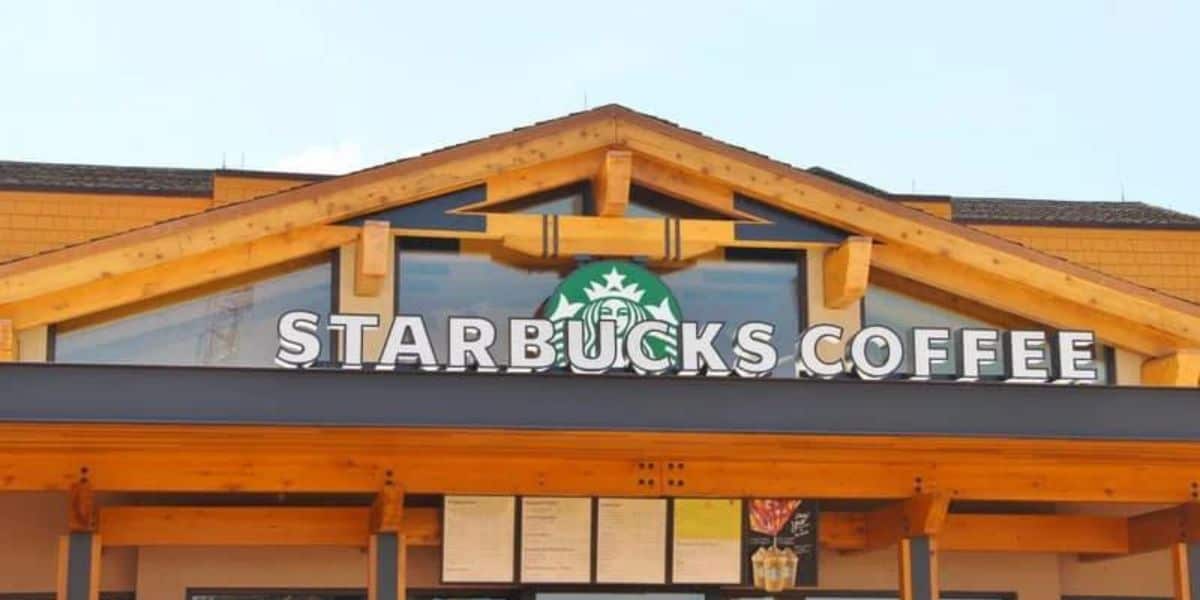 The image shows the exterior of a Starbucks Coffee shop with a wooden facade. The recognizable green Starbucks logo is above the large, white "STARBUCKS COFFEE" sign. The building has a triangular peak roof and large glass windows under the sign at Disney World.
