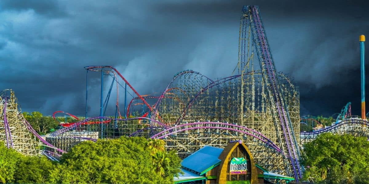 A colorful Busch Gardens Tampa Bay with numerous roller coasters, featuring twists, loops, and steep drops. The sky overhead is dark and stormy, creating a dramatic contrast with the vibrant rides. Lush green trees are in the foreground, and a building labeled "Iron" is visible.
