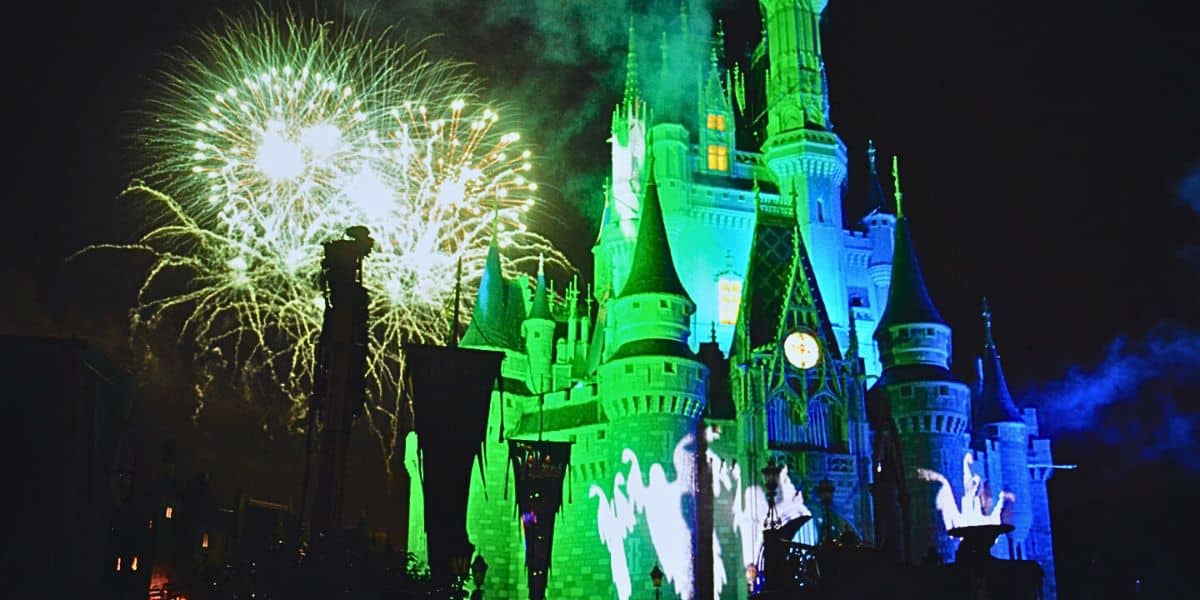 A brightly lit castle with hues of green and blue stands tall at night, illuminated by a magical light show. Fireworks burst in the sky, casting colorful reflections on the castle's turrets. Silhouettes of cheering visitors and Disney cast members are visible in the foreground.