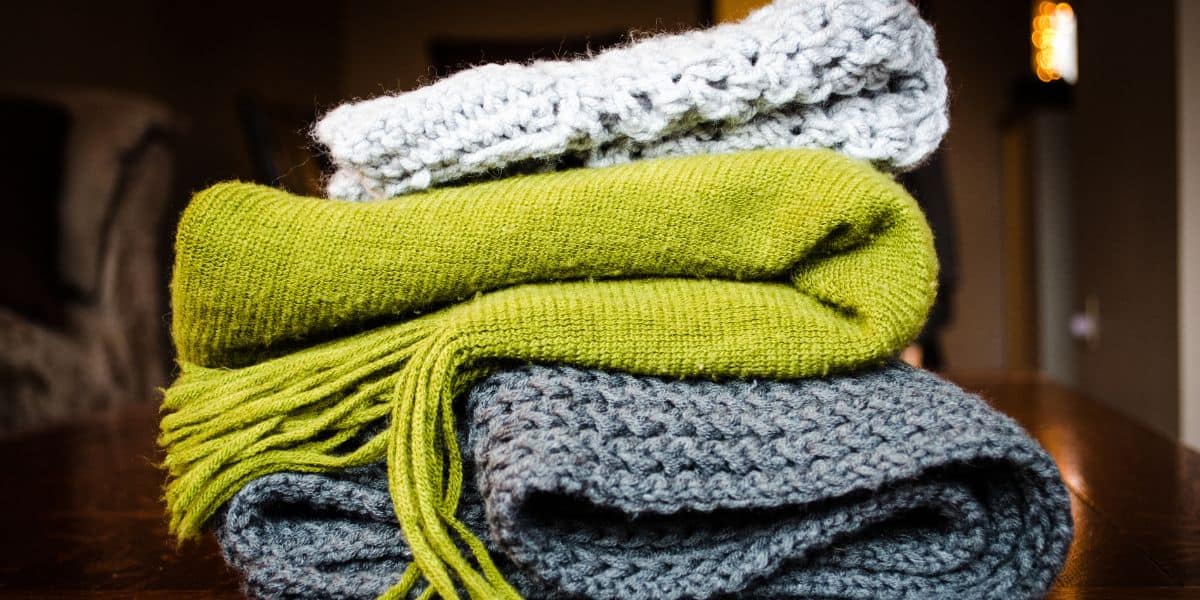 A close-up of three neatly folded knitted scarves stacked on a dark surface evokes the cozy warmth of a wilderness lodge: a white textured scarf on top, a green scarf in the middle, and a thick gray scarf on the bottom. The background is out of focus.