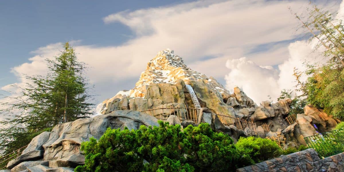 A large mountain with snow-capped peaks and rugged rocky terrain is surrounded by lush green trees and bushes. A wooden fence runs along a path near the base of the mountain, and white clouds fill the blue sky in the background at Disneyland California, with the Matterhorn in sight.