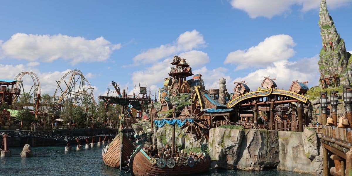 A vibrant Epic Universe theme park scene with a Viking ship on water, roller coasters in the background, rocky cliffs, fantasy architecture, and blue skies with scattered clouds.