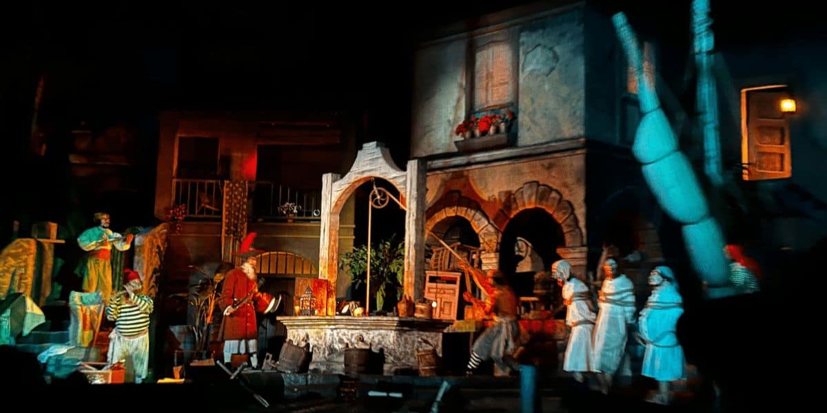 Pirates of the Caribbean, iconic Disney ride, failing during the well scene at Disneyland Park. A lively scene in a dimly lit, colorful stage set resembling a historic village. Characters in pirate and colonial costumes, reminiscent of Pirates of the Caribbean, are animatedly interacting, with a well in the center and various buildings as a backdrop.