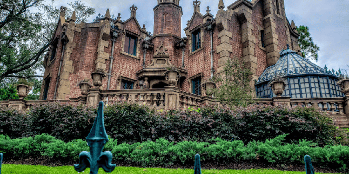 A vibrant image of the haunted mansion at Walt Disney World, showing the eerie, gothic architecture of the building surrounded by lush green landscaping under a cloudy sky.