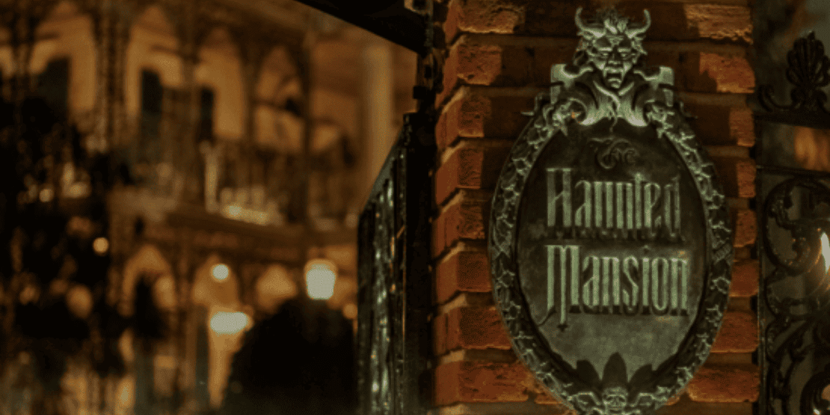 A sign that reads "Haunted Mansion" in front of the white mansion holding the attraction of the same name at Disneyland Park.