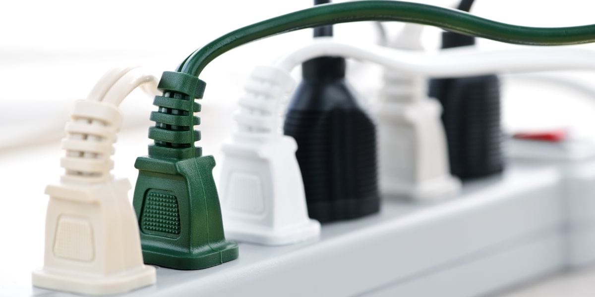 A close-up of several electrical plugs, including green, black, and white ones, plugged into a white power strip at a cozy wilderness lodge.