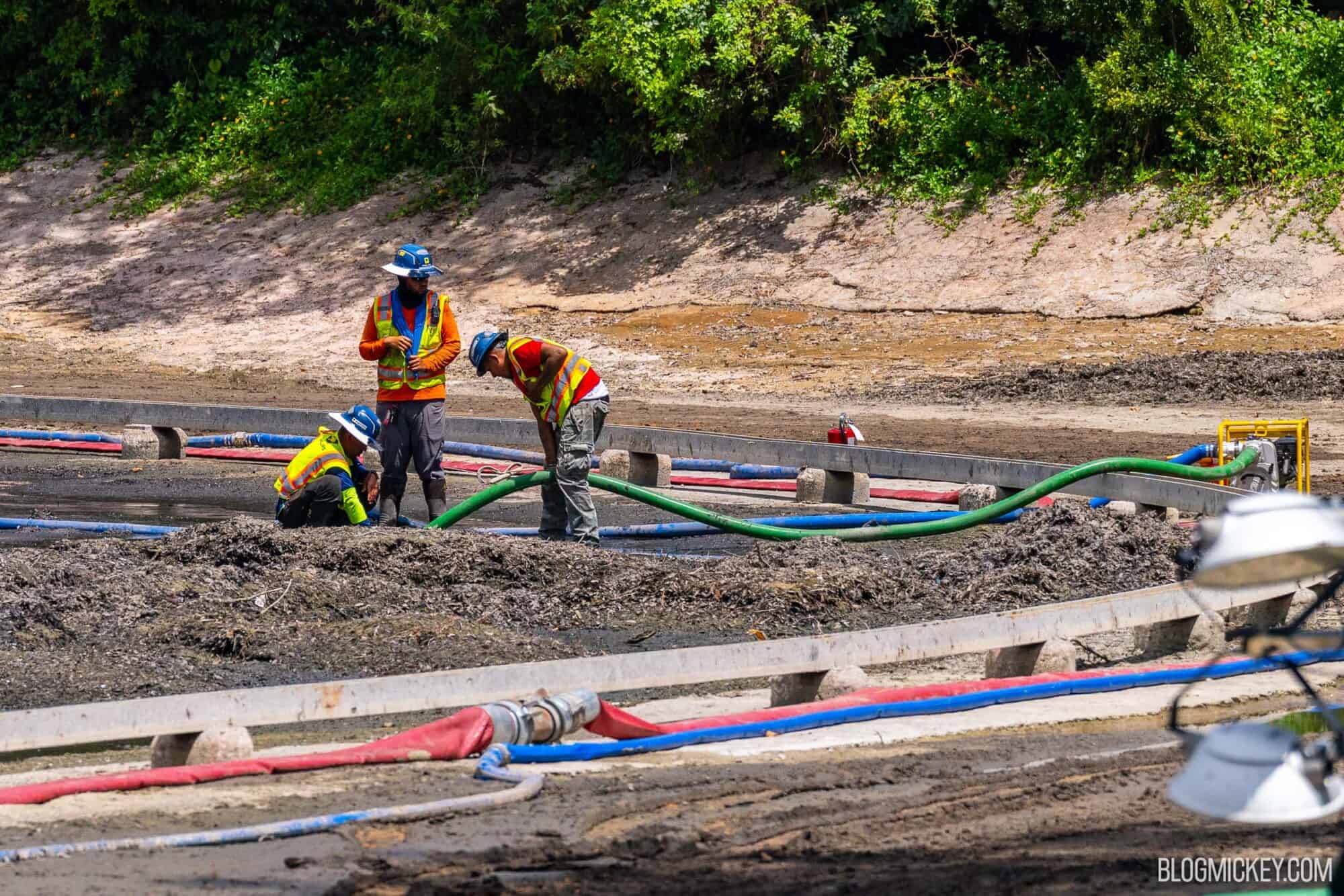 Three construction workers stand in mud, manually placing a draining hose into a small amount of water in the mostly drained Rivers of America at Magic Kingdom