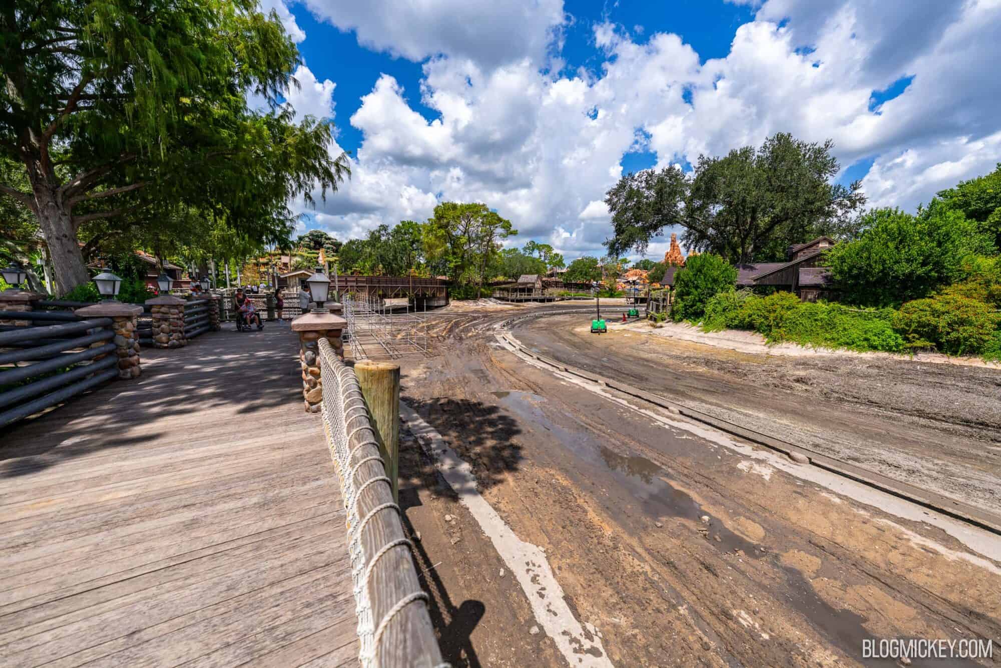 View from a wooden boardwalk of a drained Rivers of America with exposed ride track. A lush Tom Sawyer Island sits on the other side of the drained river basin
