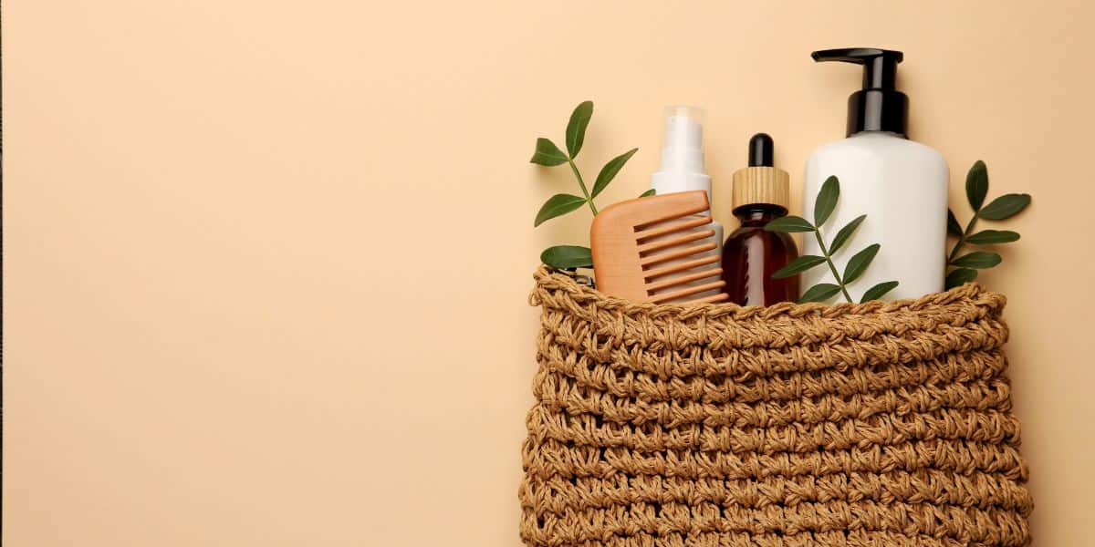 A woven basket at a wilderness lodge holds a wooden comb, spray bottle, dropper bottle, pump bottle, and green leaves, set against a beige background.