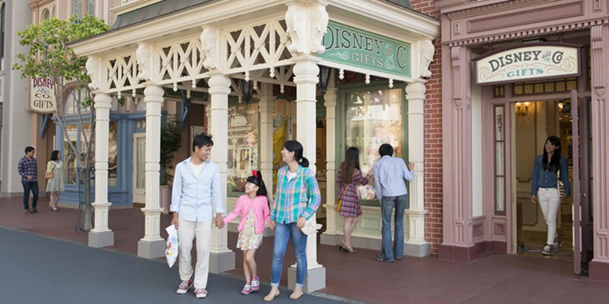A family of three, holding hands, walks past the Disney & Co. Gifts store. The father and mother are dressed casually, and their young daughter sports a pink top. The busy shopping area bustles with activity, as others queue by the storefront to explore the magical Disney merchandise inside.