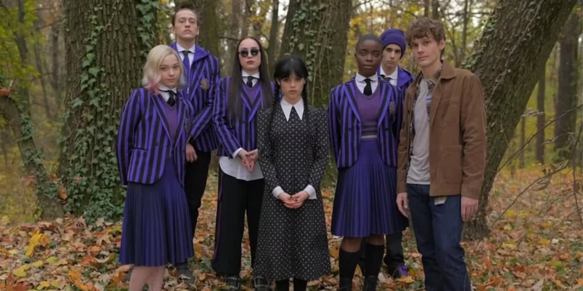 A group of seven young adults dressed in gothic-style school uniforms and casual clothing, inspired by Netflix's "Wednesday," standing in the woods, posing with a solemn expression.