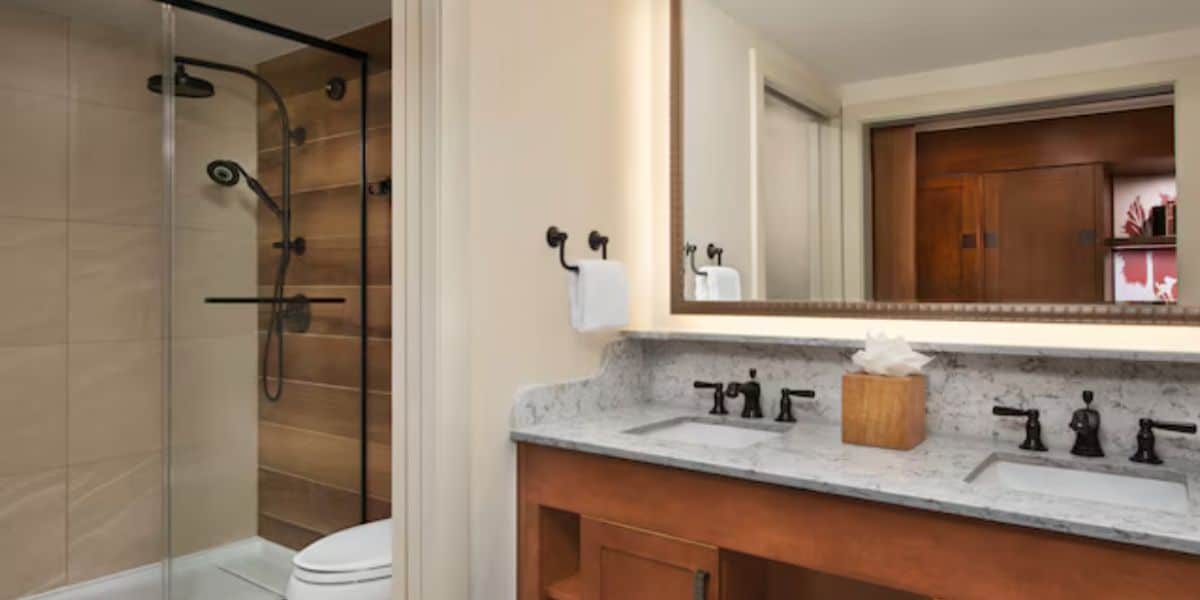 Modern bathroom with a double-sink vanity, marble countertop, large mirror, and a wooden tissue box—bringing the warmth of a wilderness lodge indoors. To the left is a glass-enclosed shower with beige tiles and a black fixture, next to a toilet.