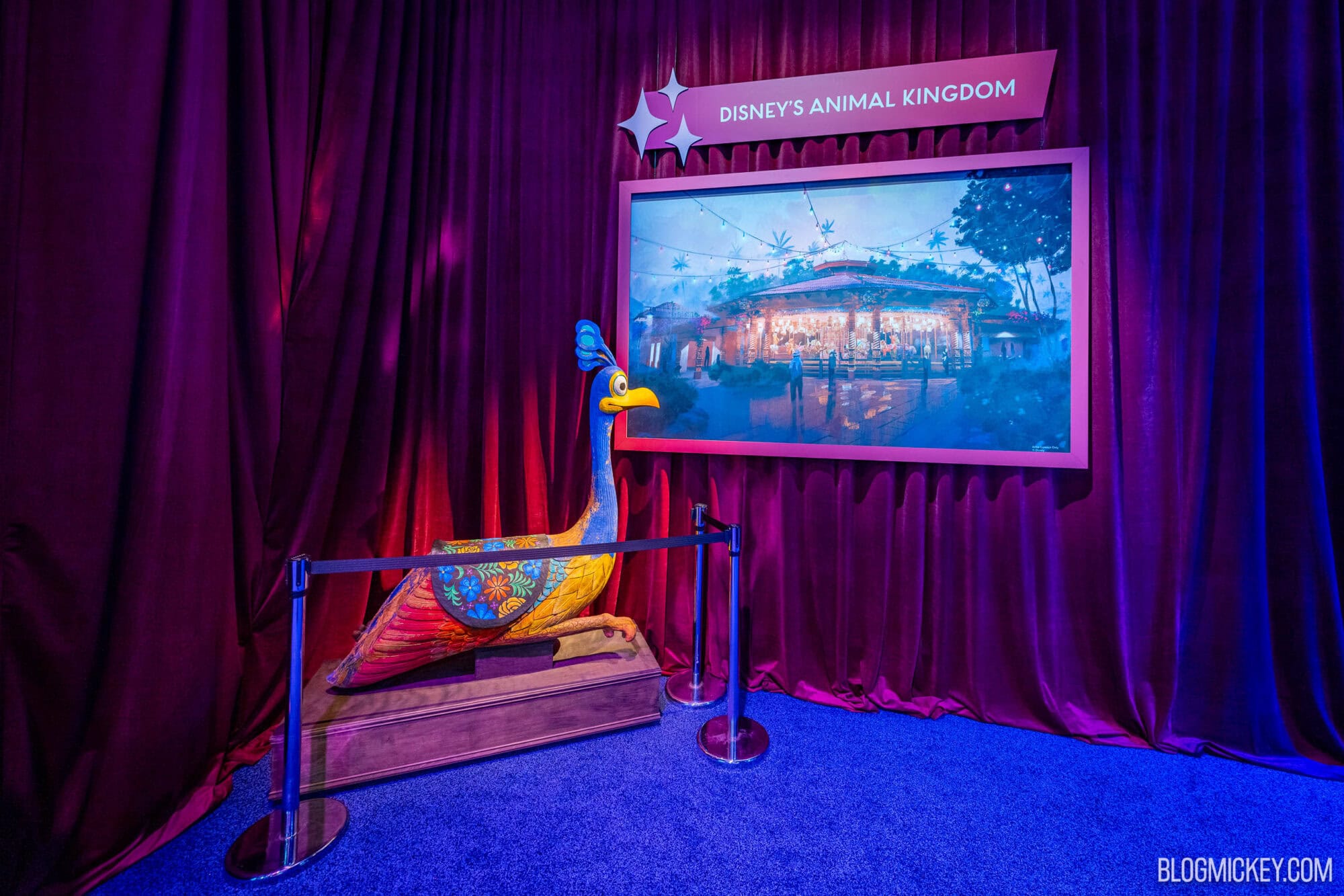 A bird-shaped ride vehicle resembling Kevin from Pixar's UP. The bird is in a seated position and made to look like it was hand carved from wood. The bird has striking blue, yellow, and red feathers. Concept art of a carousel is on a wall behind the bird