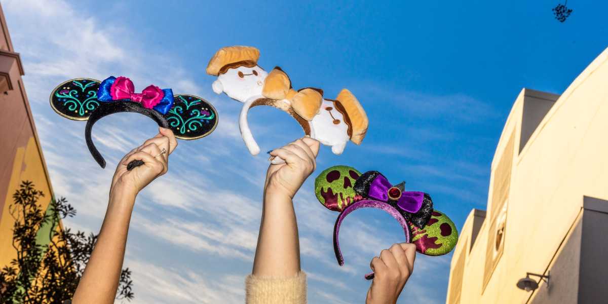 People holding up three themed headbands with Mickey Mouse ears designs against a bright blue sky, surrounded by building structures. The headbands, available at Disney’s Character Warehouse, feature various colorful decorations, including bows, intricate patterns, and character-themed designs.