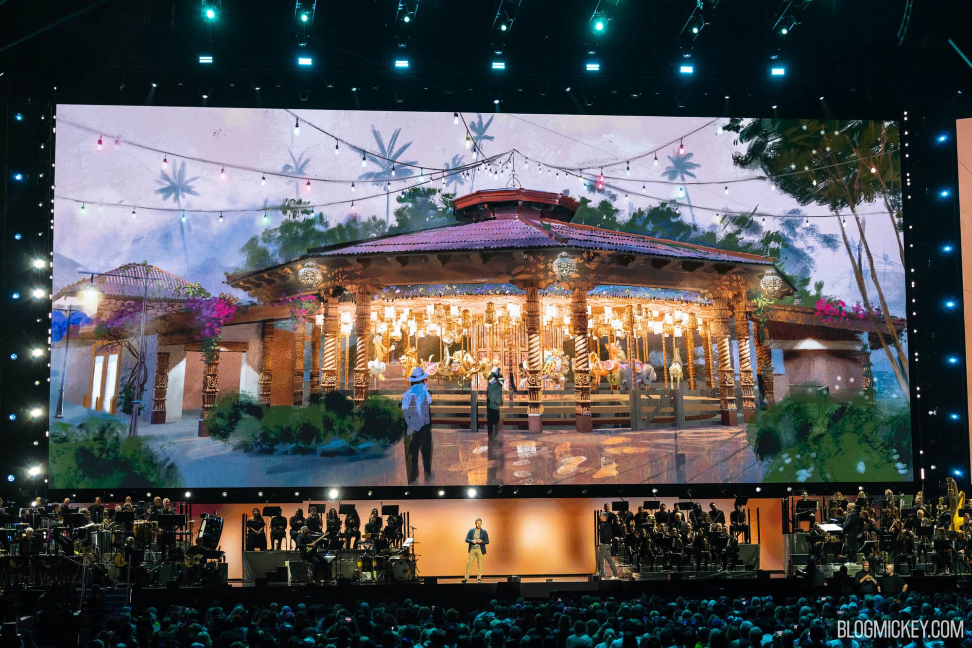 Large screen in an arena setting containing concept art of a carousel. A live band is below the screen, along with presenters