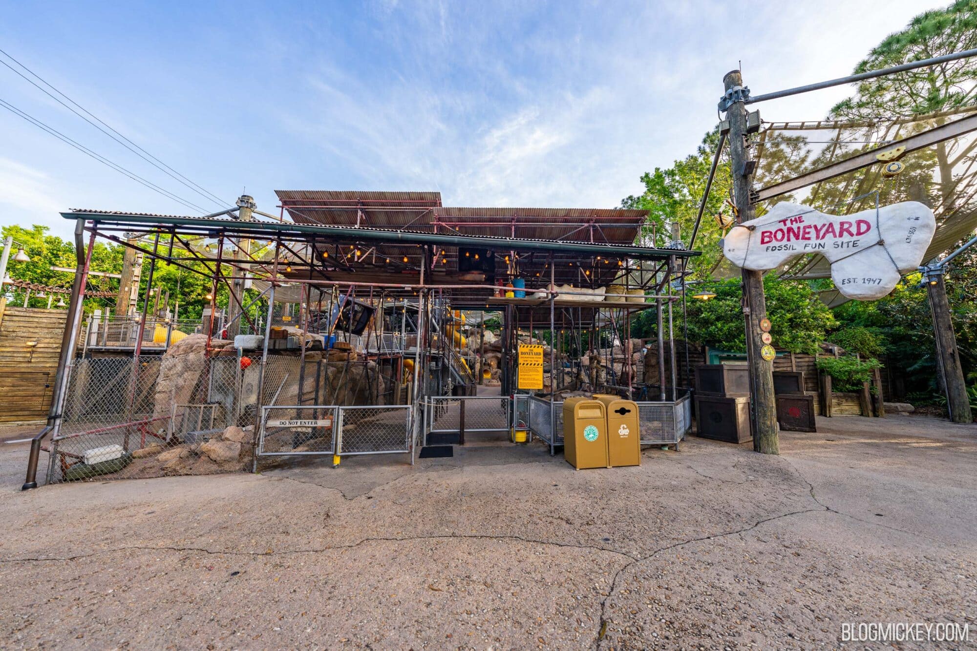 Rustic entrance to The Boneyard fossil fun site at Disney World with rocks and scaffolding, surrounded by green trees under clear blue sky, part of DinoLand USA