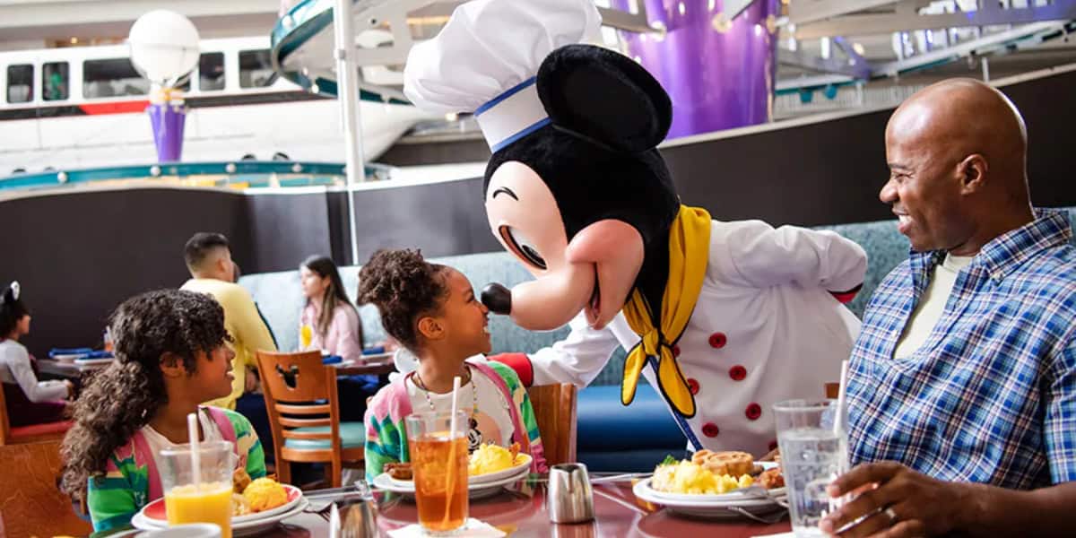 A person in a Mickey Mouse costume dressed as a chef leans down to engage with two young girls seated at a table. The table is set with plates of food and glasses of iced tea. A man, presumably their father, watches with a smile. Other diners and a monorail are in the background.