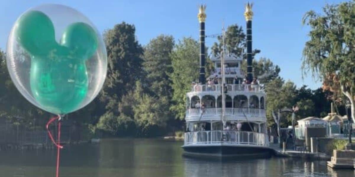A large, green Mickey Mouse-shaped balloon floats in the foreground on the left side. In the background is the Rivers of America at Disneyland Park, with the Mark Twain riverboat.