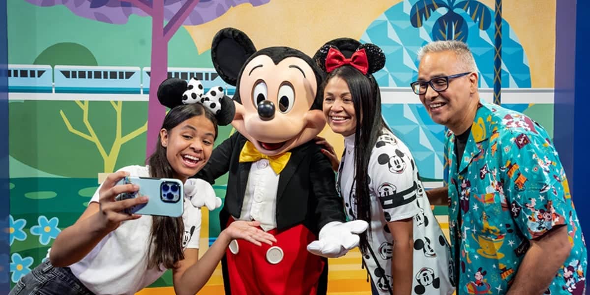 A cheerful family of three, wearing Disney-themed apparel, takes a selfie with Mickey Mouse at one of Disney's theme parks.