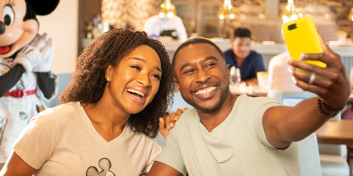 A smiling couple takes a selfie in a cheerful setting. Behind them, a person dressed as Mickey Mouse adds to the festive atmosphere. They are seated at a table, with other diners and warm lighting visible in the background.