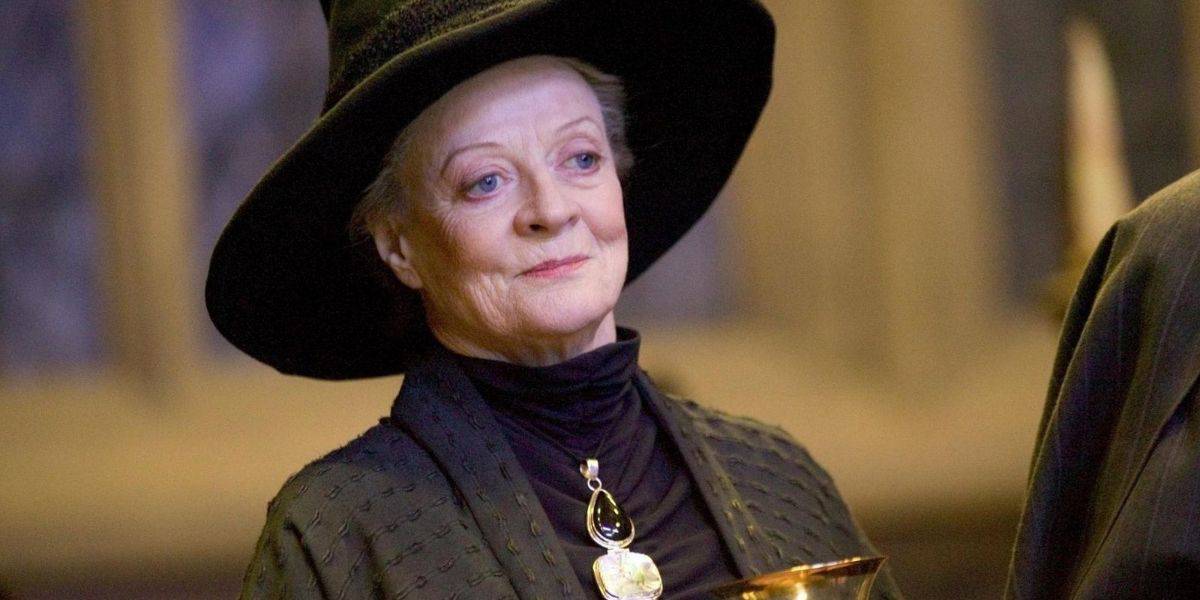 The elderly person with light skin and blue eyes, resembling Maggie Smith, is wearing a large black hat and a dark, patterned cloak. They hold a golden object and adorn a large teardrop-shaped pendant. The background suggests an indoor setting with dim lighting.