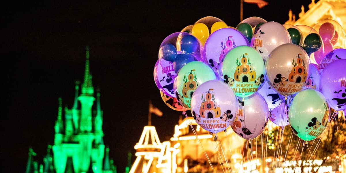 A cluster of decorative balloons featuring "Happy Halloween" messages and Disney characters floats at a night-time event. In the background, a castle is illuminated with green lights, and festive lights adorn the surroundings, adding to the Halloween ambiance.